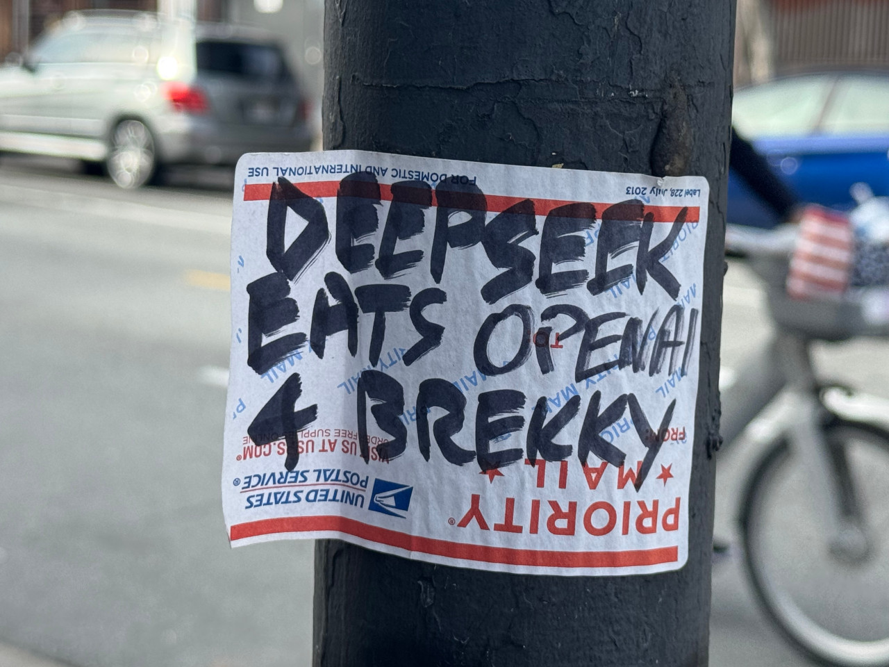 A United States Postal Service label sticker upside down on a light post with the text “DEEPSEEK EATS OPENAI 4 BREKKY” on it